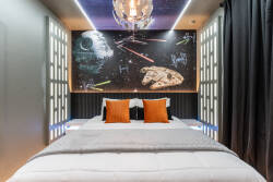 Step into a uniquely themed bedroom where a striking mural of spaceships creates a playful atmosphere. The plush bedding, accented by vibrant orange pillows, invites relaxation, while stylish lighting enhances the room’s ambiance.