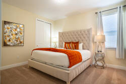 This bright, inviting bedroom boasts a plush tufted headboard and a cozy orange throw for warmth. Natural light fills the space, highlighting the soft carpet and chic accent artwork for a truly delightful stay.