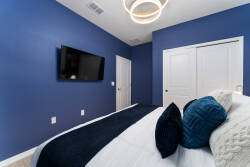 This cozy bedroom features calming blue walls, soft white bedding, and textured throw pillows for comfort. A modern light fixture and a wall-mounted TV enhance the space, making it perfect for relaxation and entertainment.