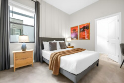 Experience a bright and airy bedroom with soft gray tones and warm wood accents, creating a serene atmosphere. Delight in vibrant wall art and stylish bedside lamps, perfect for your reading enjoyment.