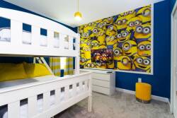 This cheerful children's bedroom boasts a white bunk bed and vibrant blue walls adorned with a fun Minions mural, complemented by a cozy yellow chair and soft carpeting, creating an inviting space for kids to play and relax.