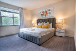 Enjoy peaceful nights in this beautifully designed bedroom with a plush gray bed and cheerful yellow pillows. Large windows let in natural light, enhancing the soft carpet and vibrant artwork that bring the space to life.