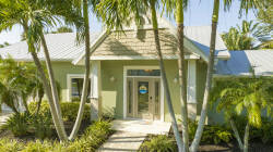 Welcome to a charming entryway framed by lush palm trees and vibrant landscaping. The light-colored façade and shingled roof offer a delightful coastal charm, setting the stage for a relaxing vacation inside.