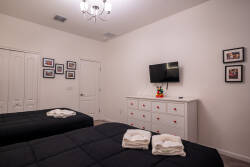 This cozy bedroom boasts two plush beds with crisp white linens and a charming red-accented dresser. Framed photos decorate the walls, and a stylish chandelier softly illuminates the space, creating a warm and inviting atmosphere.