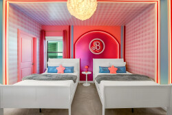 This vibrant bedroom features two stylish white beds with cozy gray throws and colorful pillows. The pink and blue walls, enhanced by glowing lights, create a lively atmosphere ideal for relaxation.