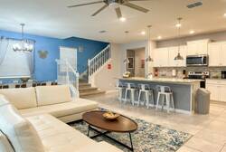 Enjoy an open living area with a cream sectional, ceiling fan, dining space, staircase, and bright kitchen with island seating, perfect for relaxing and gathering during vacation.