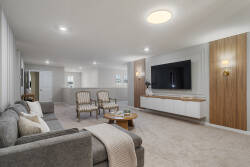 Relax in a bright and stylish living room with a cozy gray sectional, wall-mounted TV, elegant chairs, soft curtains, and warm wood accents. The neutral decor and natural light create a calm space for family vacation moments.