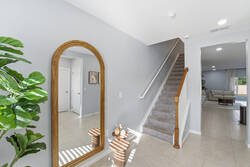 Welcome yourself into a bright entryway with a tall wood-framed mirror, soft gray walls, greenery, natural light, and a carpeted staircase, creating a clean and inviting start to your vacation.