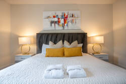 Welcome to your cozy retreat featuring a king-sized bed with a plush tufted headboard, crisp white linens, and vibrant yellow pillows. Stylish bedside lamps illuminate contemporary artwork, creating a warm and inviting ambiance.