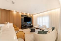 Relax in the bright, airy living area with soft white furnishings and warm wood accents for a cozy feel. The sleek black entertainment unit adds sophistication, while large windows fill the space with inviting natural light.