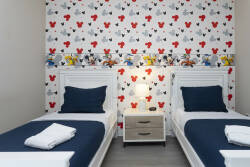 This cozy bedroom welcomes you with two twin beds dressed in crisp white linens and navy blue accents, alongside whimsical wallpaper featuring colorful characters. A soft bedside lamp casts a warm glow, perfect for restful nights.