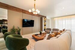 Relax in a bright open living area with a large sofa, green accent chairs, wall-mounted TV, modern lighting, and a nearby kitchen, creating a comfortable space to gather during your vacation.