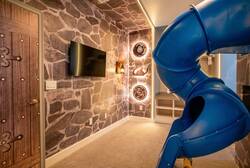 Welcome to a whimsical playroom with a vibrant blue slide and a charming stone-patterned wall. The cozy carpet and unique decor make it the perfect space for family fun and relaxation together. Enjoy your time!
