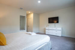 This bright, spacious bedroom offers a relaxing retreat with plush carpet, crisp white bedding, and a cheerful yellow pillow. Enjoy the convenience of a sleek dresser and wall-mounted TV during your stay.