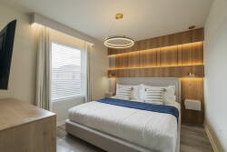 This stylish bedroom boasts a plush bed with crisp white linens and deep blue accents, warm wood details, and soft lighting, while the inviting layout and large window create a refreshing space perfect for relaxation.