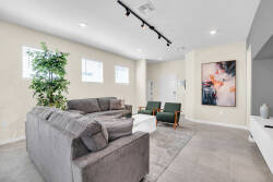 This bright and airy living area features a plush gray sectional for relaxation, stylish green armchairs, and artistic wall art lit by track lighting, while large windows fill the space with natural light for a warm atmosphere.