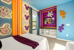 This vibrant bedroom, adorned with whimsical art and cheerful accents, invites relaxation. The cozy bed, featuring a deep violet throw, welcomes you as natural light streams through the large window, brightening the lively decor.