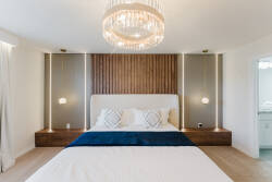Discover a beautifully designed bedroom with a stunning chandelier above a plush king-sized bed dressed in crisp white linens and a rich blue throw, while warm wooden walls and sleek nightstands add to the modern comfort.