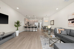 Welcome to a bright living room with a sleek gray sectional and stylish coffee table, perfect for relaxation. The open layout flows into a chic kitchen with modern bar stools and warm lighting, creating an inviting space for your.
