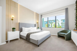 Experience a bright and airy bedroom with a plush king-sized bed dressed in crisp white linens, soft beige walls, and chic sconces. Enjoy natural light streaming through large windows and a cozy accent chair for added comfort.